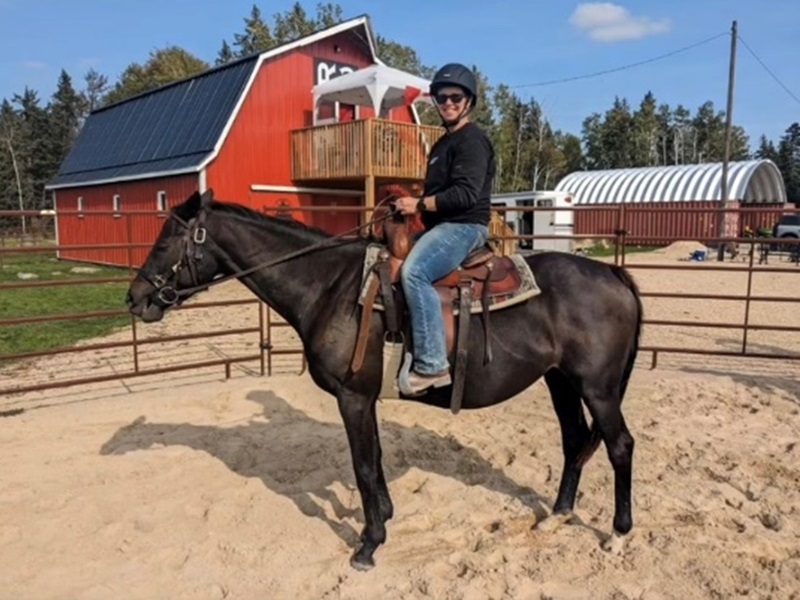 A woman riding a horse in a fenced outdoor arena at Red Rose Ranch, with a red barn and trees in the background.