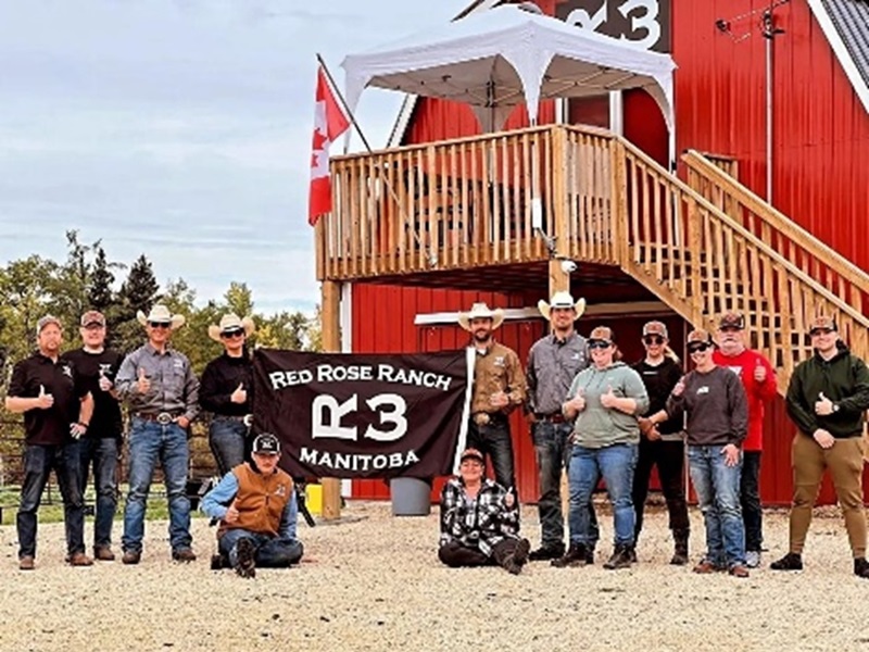 A group of people in western-style clothing pose with a “Red Rose Ranch Manitoba” banner in front of a red barn with a Canadian flag.