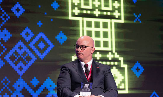 Brigadier-General Scott MacGregor listens attentively during a panel discussion at CyberChess 2025 in Riga, Latvia, with a digital chess rook displayed on the screen behind him.