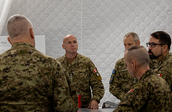 Five members of the Canadian Armed Forces wearing uniforms stand around a map table having a conversation in a large tent.