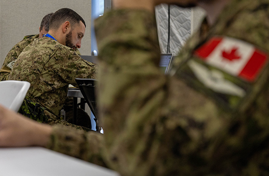 Three uniformed members of the Canadian Armed Forces sit at computer workstations in a large tent, the member in the foreground is out of focus and the two in the background are in focus.