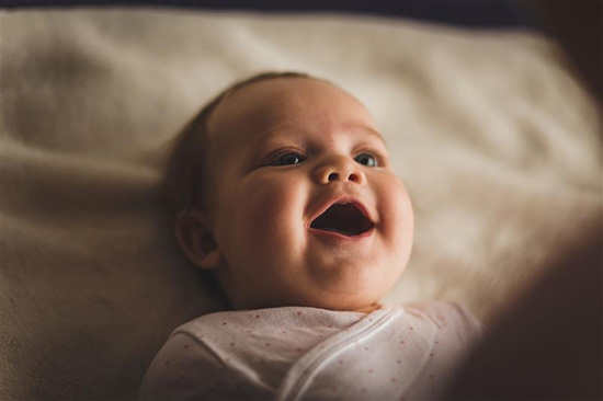 Baby with blue eyes, wearing a pink polka dot shirt, lying down on a sheet, and looking up with mouth open.