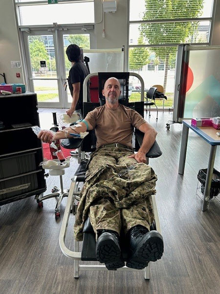 A military member sitting in a long chair, smiling at the camera while donating blood.