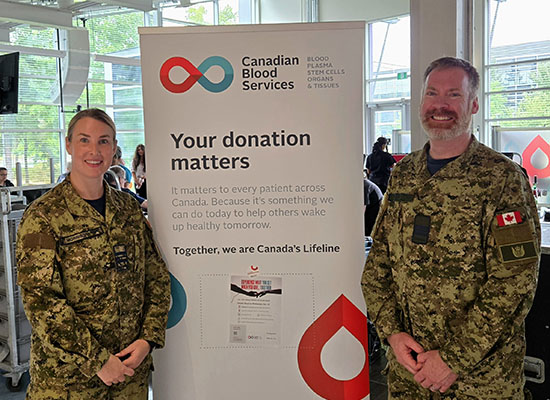 Two military members standing on each side of a blood drive’s main pop-up display and smiling at the camera.
