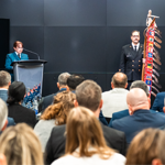 An audience is seated facing a podium where a speaker is addressing the group. Beside the podium stands a uniformed individual next to a ceremonial staff adorned with colorful ribbons and feathers. The setting appears to be an indoor conference or formal event, with cameras positioned to record the proceedings.
