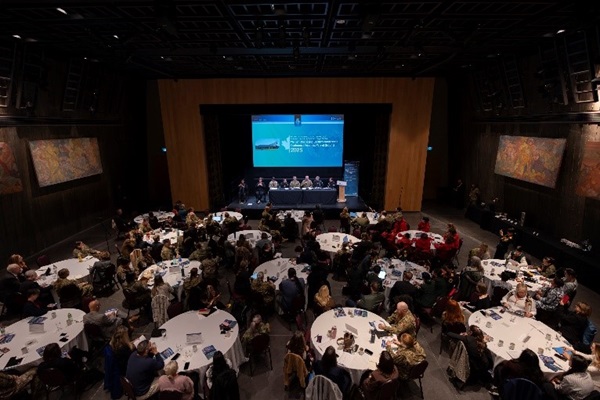 Image d’une conférence avec plusieurs tables rondes entourées de participants. Un écran de projection affichant une présentation se trouve à l’avant de la salle avec une table de panélistes au-dessous.