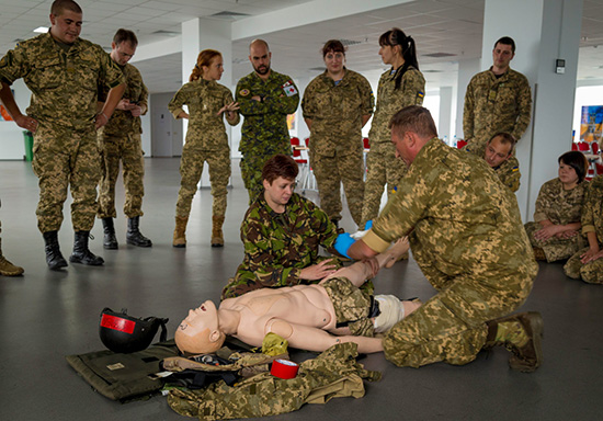 A soldier in uniform watches closely as other soldiers administer care to a simulated casualty.