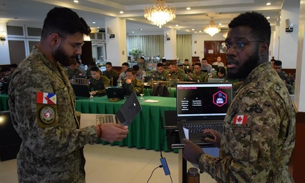 Deux cyberinstructeurs des Forces armées canadiennes coordonnent leurs efforts sur un ordinateur portable tandis que le personnel des Forces armées des Philippines suit une formation pratique sur la cybersécurité dans une salle de conférence.