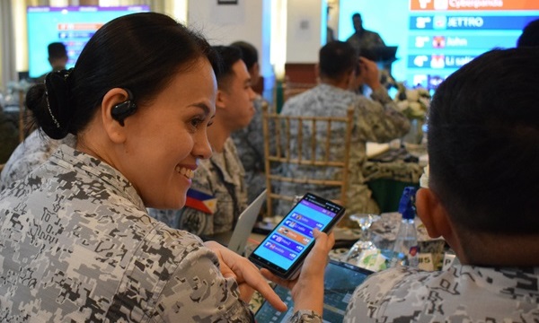 Un participant des Forces armées des Philippines examine l'interface d'un cyberexercice sur un téléphone intelligent, assis parmi ses collègues pendant une session de formation.