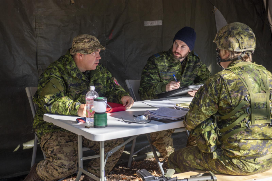 Three soldiers in uniform sit around a folding table inside a tent, reviewing documents and taking notes as part of an outdoor military exercise. 