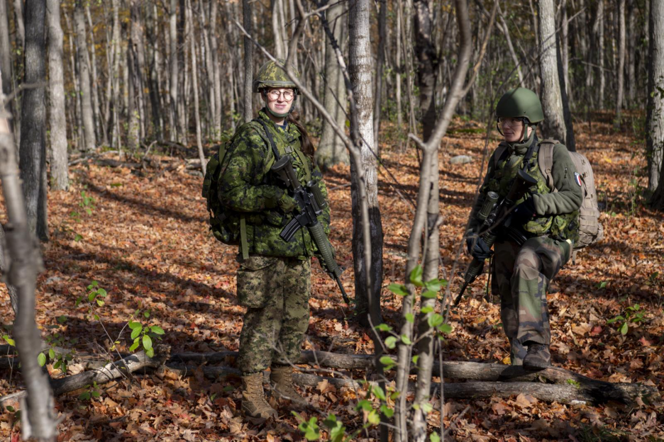 Two legal officers in uniform stand in a forest during autumn training, surrounded by fallen leaves. 
