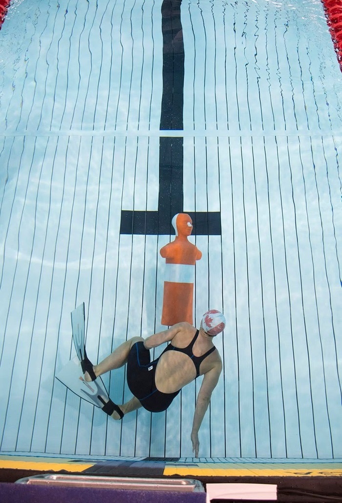 A swimmer wearing fins, a swim cap, and a dark one piece suit is underwater in a swimming pool, positioned horizontally near the pool floor. The swimmer is reaching toward an orange life saving training mannequin that is resting on the bottom lane line.