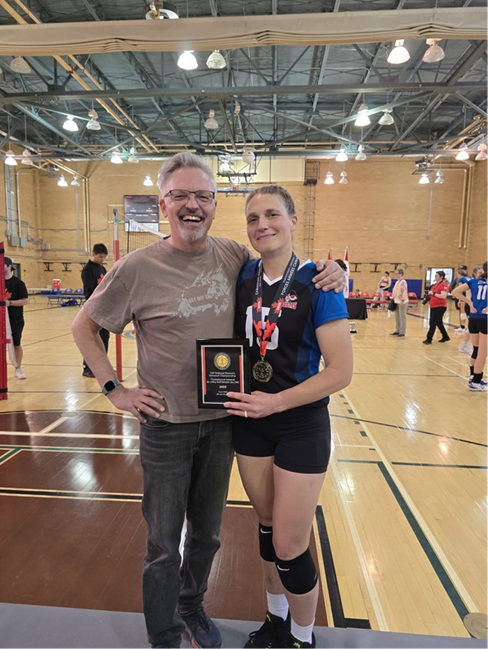 Two people stand together. One person is wearing athletic gear with knee pads and a medal around their neck while holding a plaque. The other person is standing beside them with an arm around their shoulder. Volleyball nets, players, and gym equipment are visible in the background.