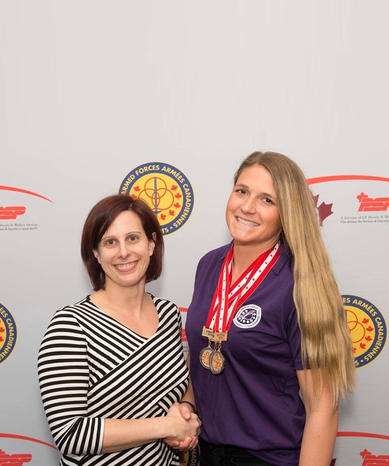 Two individuals shaking hands in front of a backdrop featuring Canadian Armed Forces sports logo. One individual is wearing a striped long sleeve shirt, and the other is wearing a purple shirt with several medals hanging from red-and-white ribbons around the neck.