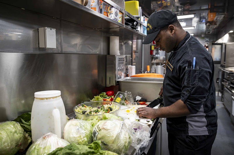Un militaire vêtu de noir coupe des aliments pour une salade dans une cuisine.