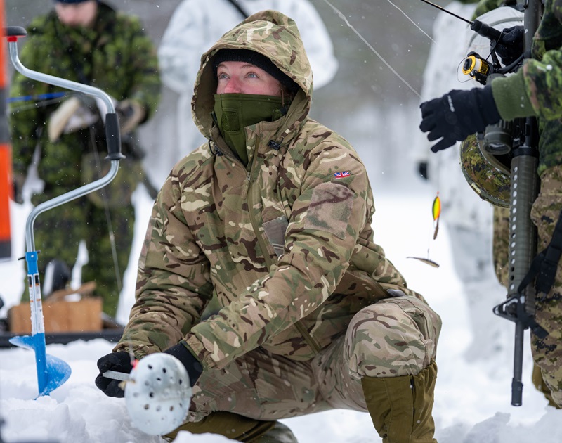 UK military nurse kneeling on snow while drilling and preparing an ice-fishing hole during training.