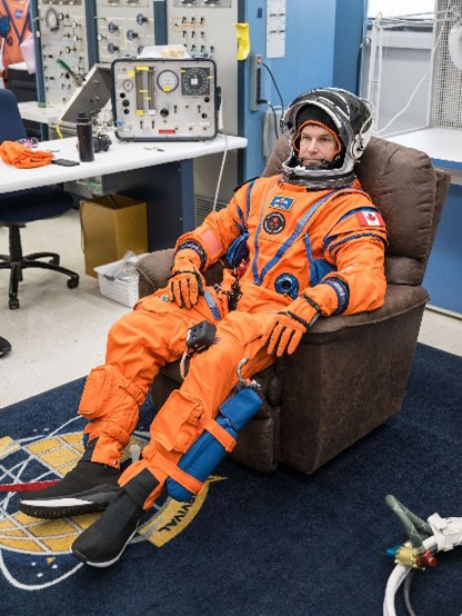 A man wearing a custom-built bright orange astronaut flight suit and a white and black astronaut helmet, participates in the testing of the Orion Crew Survival System for safety.