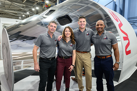Four people, three men and one woman, stand in front of a NASA Space Launch System crew capsule. They stand close together with their hands on each other’s shoulders.