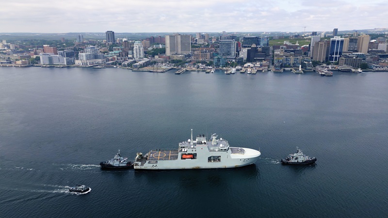 Large naval vessel on the water escorted by smaller boats, with a city skyline in the background.