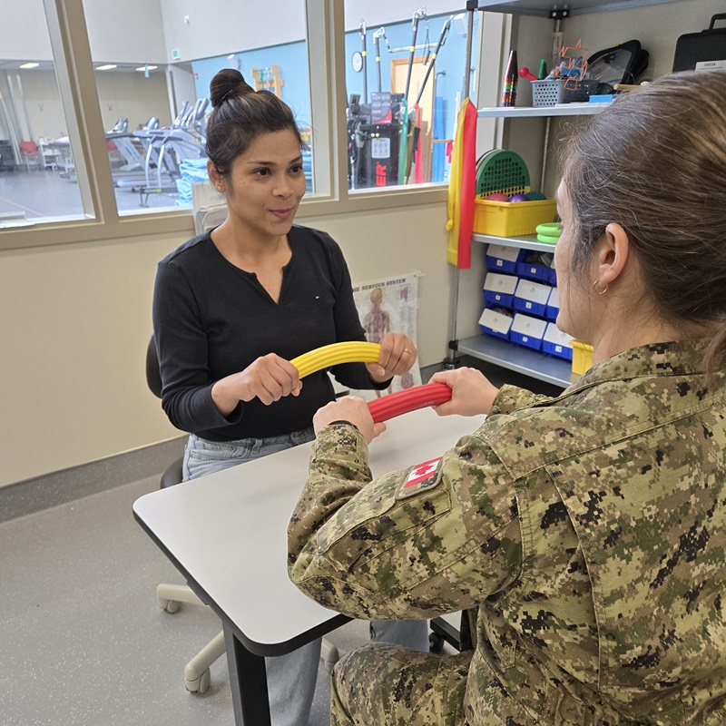Occupational therapist guides a uniformed member through grip strength exercises using resistance bars.