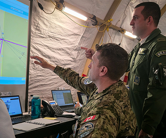 Canadian and Brazilian military personnel in a command center analyzing a large digital map displayed on a screen during strategic planning session.