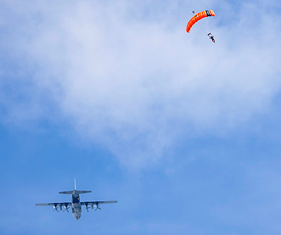Royal Canadian Air Force CC-130H Hercules aircraft flying in a blue sky with a parachutist descending under an orange parachute.