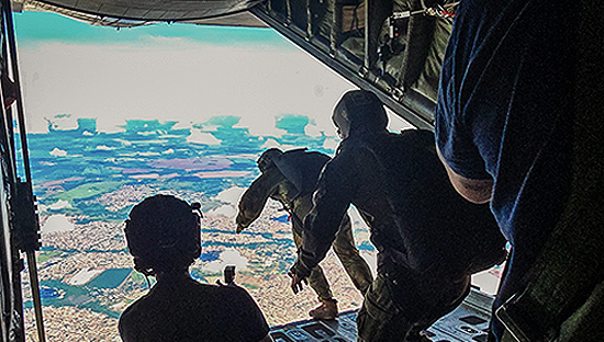 Three Brazilian military members inside a Royal Canadian Air Force CC-130H aircraft prepare for a parachute jump, with one person positioned at the open rear door ready to exit.
