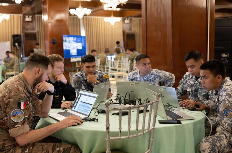 A CAF cyber expert and allied military personnel sit around a table using laptops, collaborating during a multinational cyber defence exercise indoors.