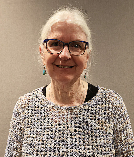Picture of a woman wearing a white shirt with holes and she is smiling and has white hair and black glasses.