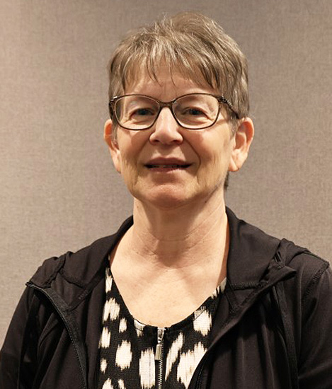 Picture of a woman with black glasses, she has short grey hair and is wearing a black and white blouse and black vest.
