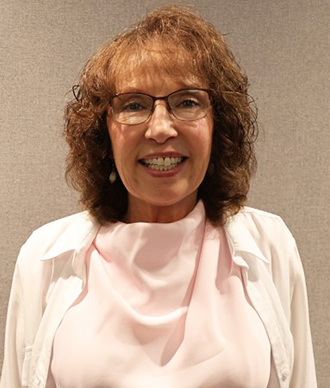 Picture of a woman wearing a pick blouse and white vest. She has auburn hair and has glasses and is smiling.