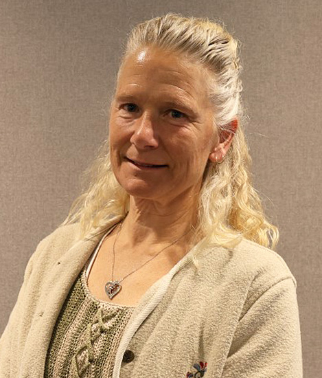 Picture of a woman who has white blond hair who is wearing a beige blouse and shirt is smiling in front of a grey wall with a frame on the wall.