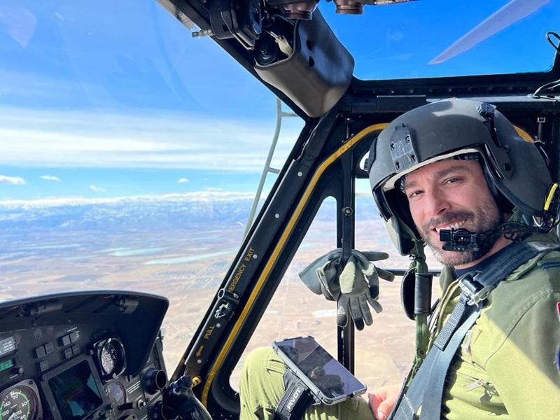 A soldier is flying a helicopter over land and smiling at the camera.