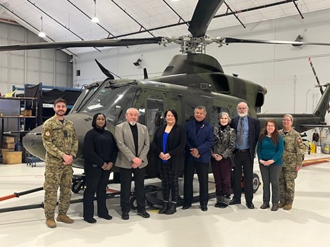 A group of ten individuals stands in a line inside an aircraft hangar in front of a military helicopter. Some individuals wear Canadian Armed Forces uniforms, while others wear civilian business attire. The helicopter is olive green with its rotor blades visible above. Maintenance equipment and hangar structures appear in the background.
