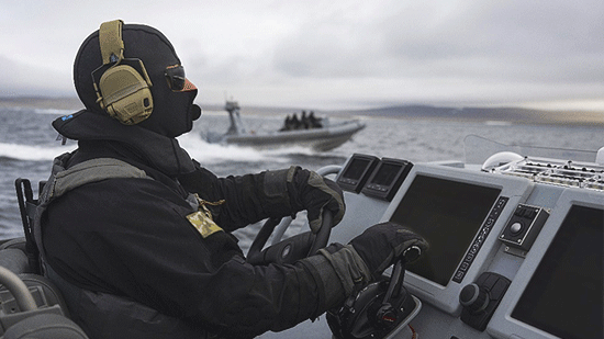 An individual wearing a black winter jacket, balaclava, sunglasses and gray load bearing life jacket is sitting at the controls of a small boat moving a high speed in the sea under a cloudy sky, while another small gray boat is visible but blurry in the background, with four similarly dressed individuals. A shore of low hills can be seen further in the background.