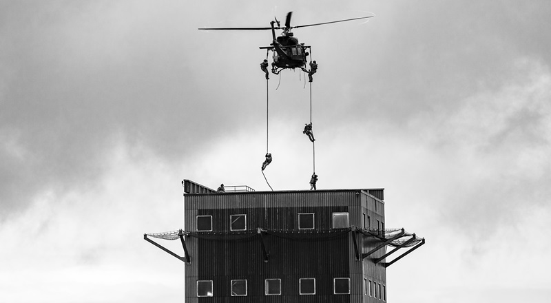 A helicopter hovers above a building that is surrounded by safety nets, under an overcast gray sky. Ropes are dangling from both sides of the helicopter and two individuals are seen sliding down on the left side rope, while three are sliding down the right side one.