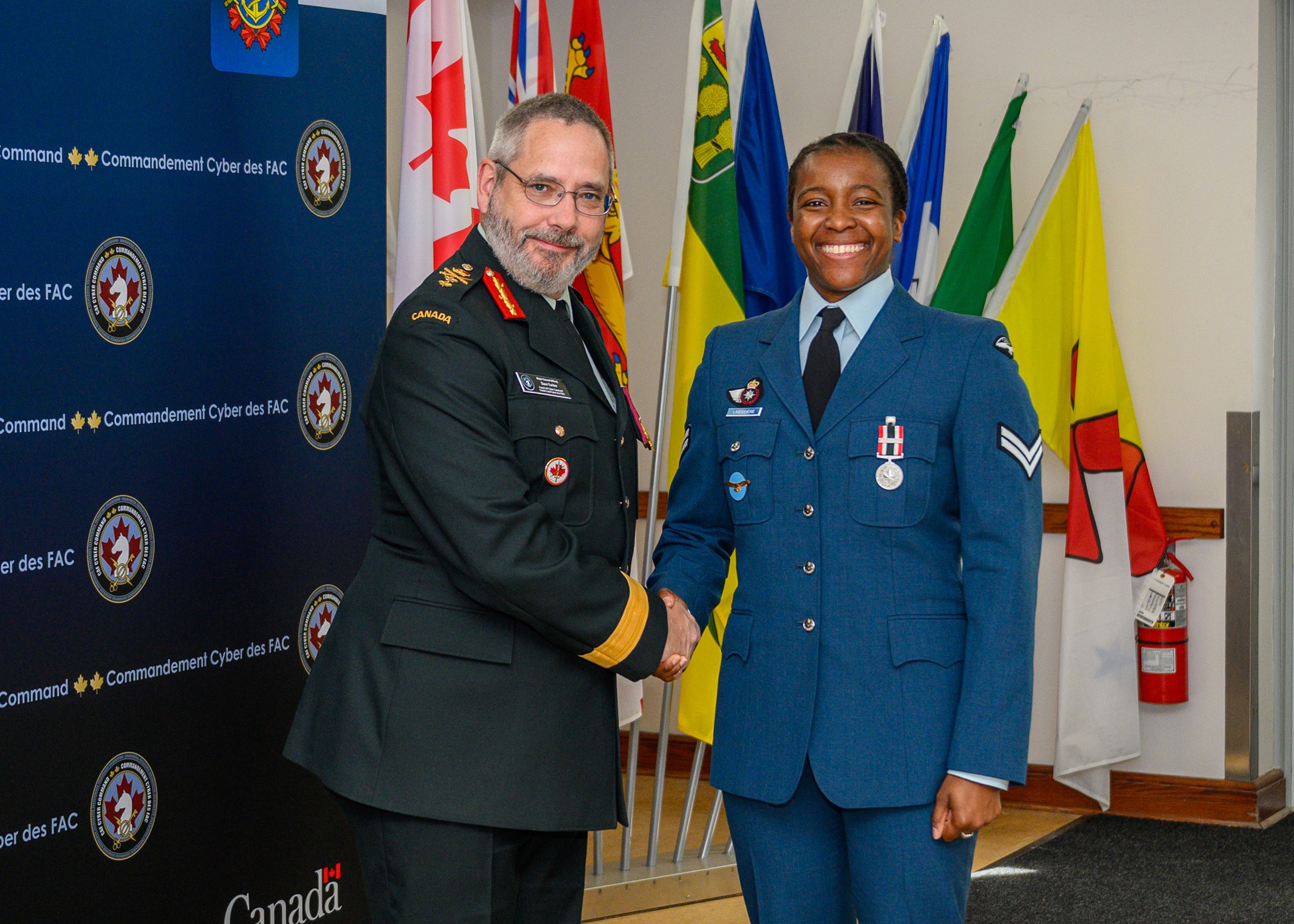 Two Canadian Armed Forces members in uniform shake hands during an indoor ceremony, with flags and a Canadian Armed Forces Cyber Command backdrop behind them. The person on the right wears a Special Service Medal with the “DISTANTIA” bar on the chest.
