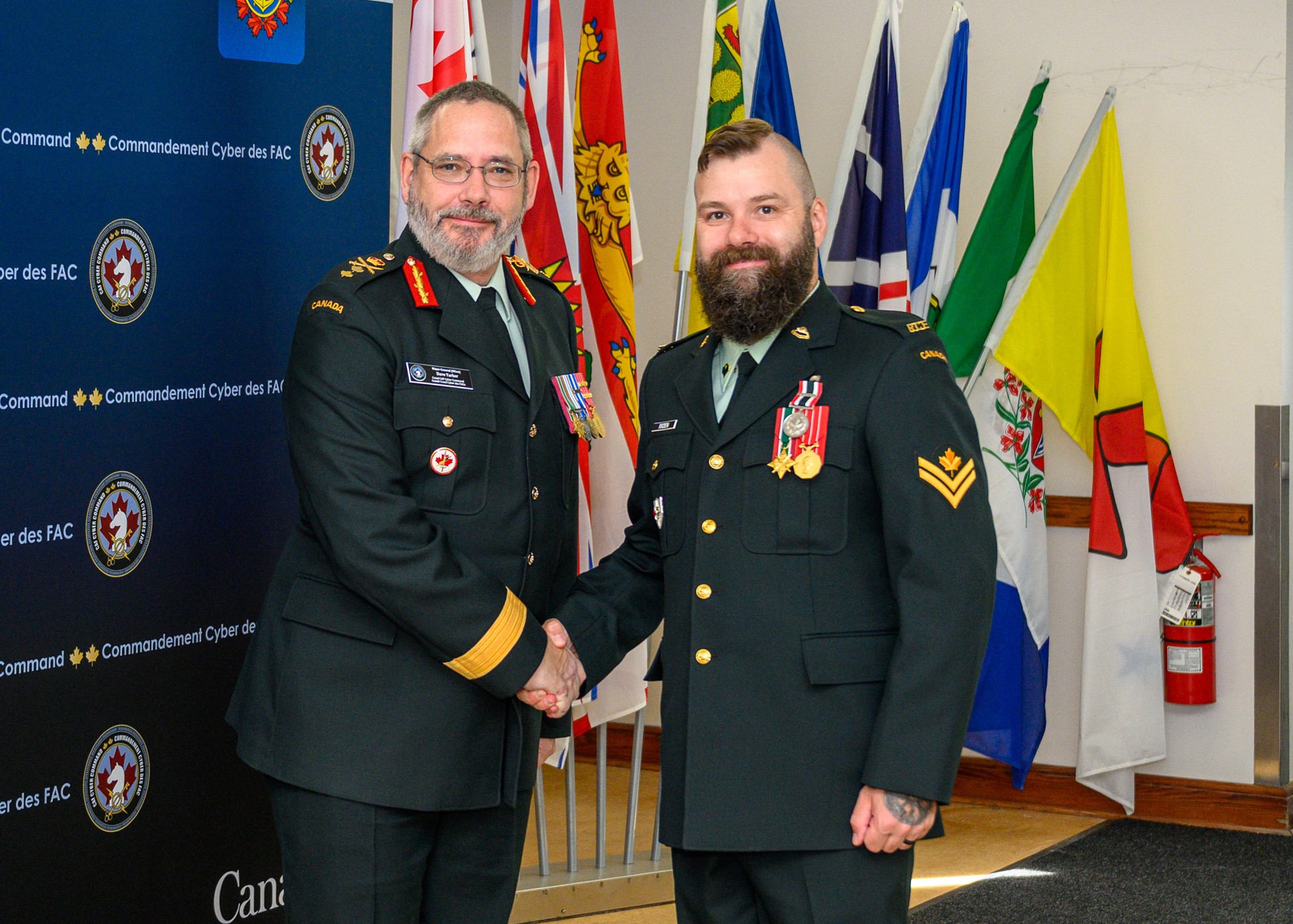 Two Canadian Armed Forces members in uniform shake hands during an indoor ceremony, with provincial and national flags and a Canadian Armed Forces Cyber Command backdrop in the background. The member on the right wears several medals, including a Special Service Medal with the “DISTANTIA” bar displayed on the chest.