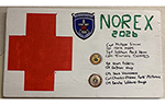 A hand-painted NOREX 2026 commemorative board featuring a large red cross, unit patches, challenge coins, and the names of Canadian Armed Forces medical personnel who participated in the exercise.