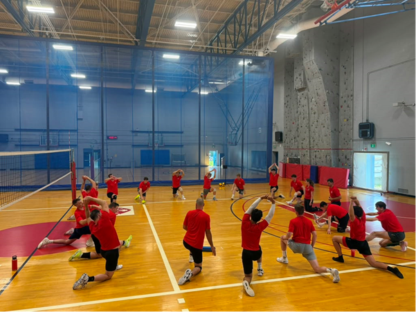 A group of athletes doing a mobility activity in a circle inside a bright indoor volleyball training facility.