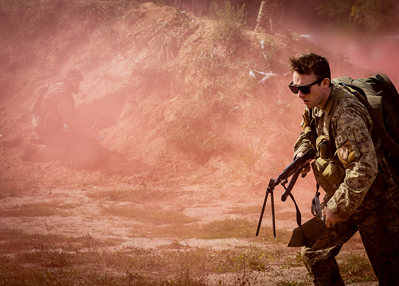 Camouflaged solider moves through drifting red smoke in a dusty field.