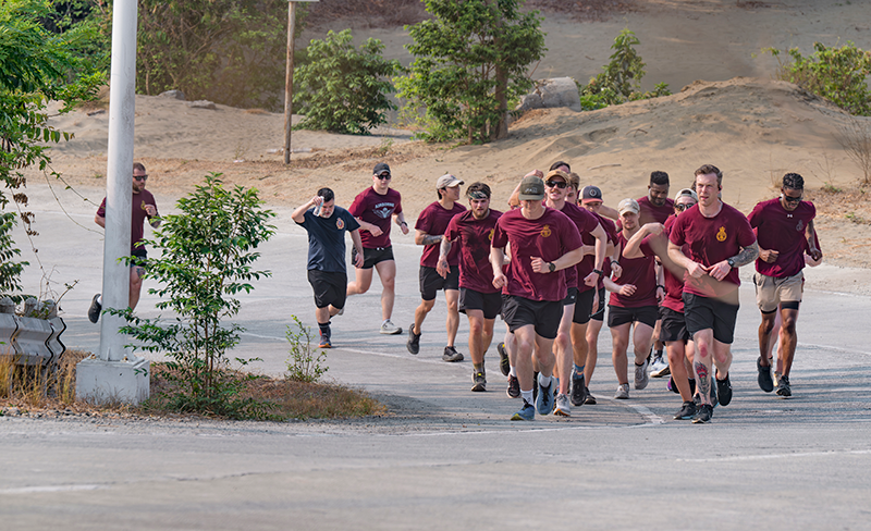Group of runners in maroon shirts run along a sunlit road in a dry, sandy landscape with sparse trees.