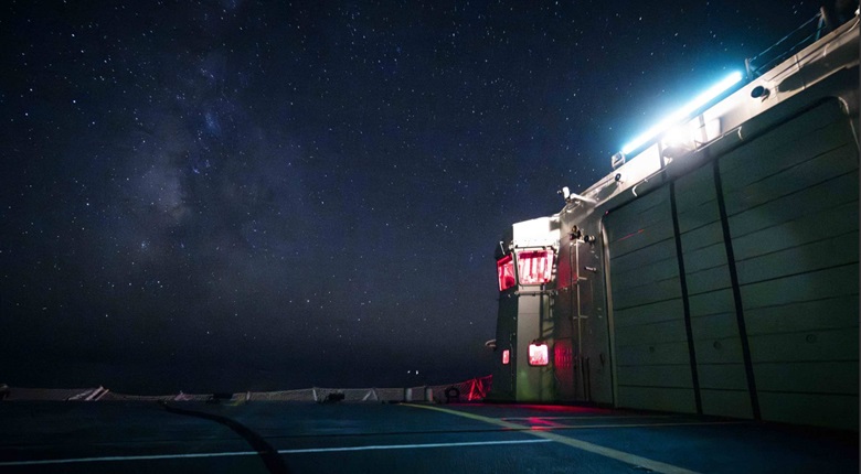 Exterior view of a large industrial building at night under a star-filled sky, with bright exterior lighting and a shoreline visible nearby.