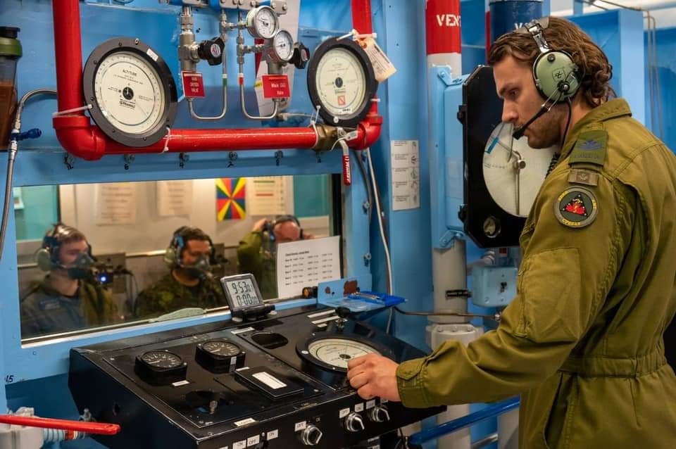 Military member uses gauges and controls to run decompression simulation in chamber, trainees visible through window.