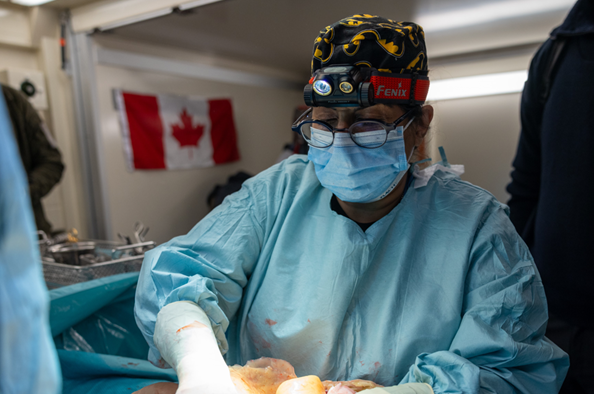 Masked clinician in gown and headlamp performs simulated training surgery, Canadian flag in background.