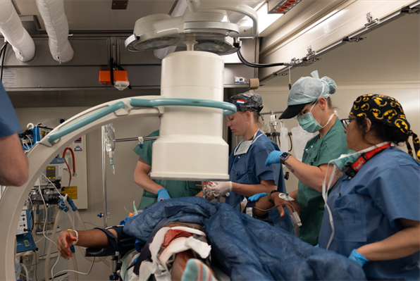 Medical team in operating room performs simulated surgical training on mannequin beneath large imaging equipment.