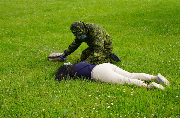 Camouflaged trainee in gas mask kneels on grass, performing simulated first aid on practice casualty during field exercise.