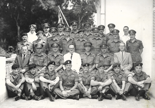 Black-and-white historical group photograph showing approximately 30 military personnel and several civilians posed on the steps and veranda of a building. Most of the military members wear service uniforms with berets or peaked caps, while a few civilians wear suits or light-coloured shirts. The group is arranged in several rows, with some seated on the ground in the front and others standing behind.