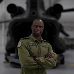 Cdr Ajiri Ikede, a Canadian Armed Forces medical officer in a green flight suit stands with arms crossed in front of a blurred CH-147F Chinook helicopter.