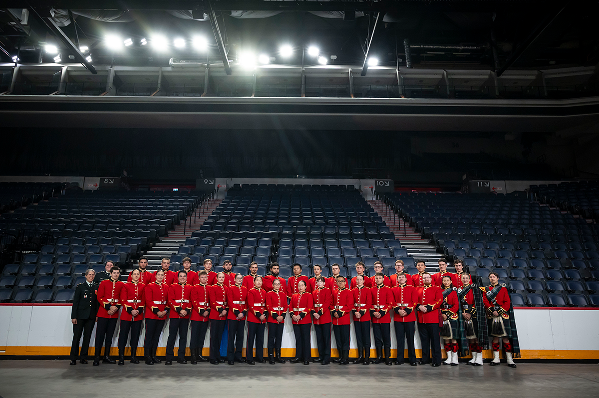 Thirty-five Royal Military College of Canada cadets wearing their ceremonial dress uniform (scarlets) and two supervising officers posing in two rows for a group photo on the grey, concrete performance floor of the Scotiabank Centre in Halifax, Nova Scotia. Bright white spotlights can be seen in the rafters.